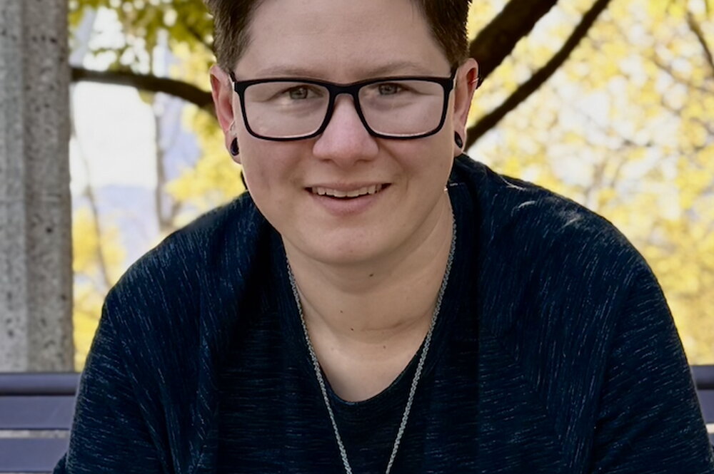 A person wearing glasses in a dark shirt standing in front of a tree